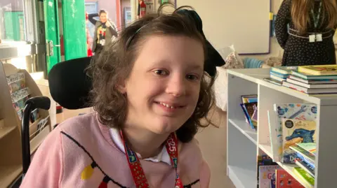 Lana is in a classroom. She is wearing a pink jumper, she has a red lanyard around her neck. She has shoulder length brown hair. There is a shelf of books to her right hand side. And an open green door behind her with a person standing in the entry. 