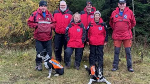 A group of mountain rescue volunteers wearing red jackets and waterproofs, four men and two women, all looking at the camera, with two collie dogs wearing fluorescent orange coats.