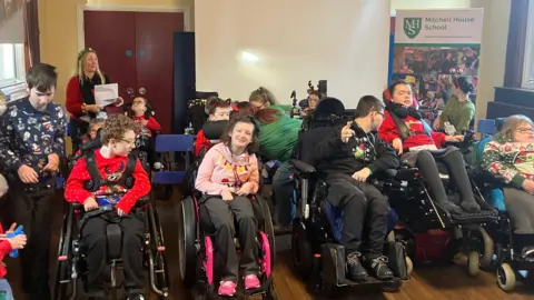 Children in wheelchairs are sitting in two lines in front of a poster. There is a woman behind them and double brown doors. 