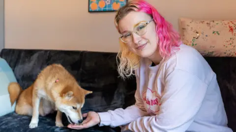 Woodgreen Ciara Pollen is smiling at the camera while Fern the dog eats treats from her outstretched hand. Ciara has blond and pink wavy, shoulder-length hair and is wearing a pale pink sweatshirt. She and the dog are sitting on a sofa in a living room.